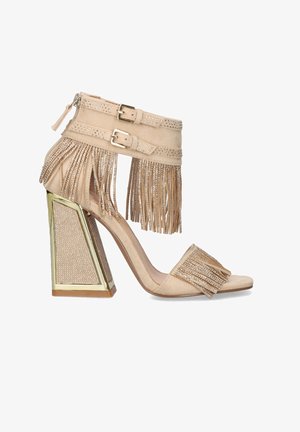 Sandalia beige de tacón alto con tacón block dorado, tiras de ante con flecos decoradas con pequeñas piedras de strass, y cierres de doble hebilla en el tobillo.