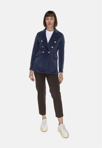 Blazer doppiopetto blu navy con bottoni dorati, design strutturato, due tasche frontali, indossato su una camicia bianca e abbinato a pantaloni marroni.