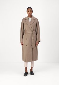 Un manteau croisé ceinturé en laine marron clair, dot�é de grands boutons et d'un col cranté, avec des manches larges et une coupe à mi-genoux.