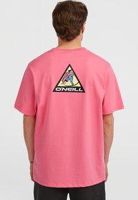 Hombre que lleva una camiseta rosa con un logo de triángulo bordado en negro que muestra a un surfista y el texto "O'Neill" en la parte posterior, sobre un fondo liso.