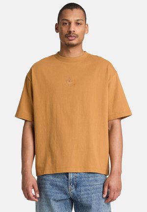 Mann trägt ein oversized senfgelbes Timberland-T-Shirt und hellblaue Jeans und steht vor weißem Hintergrund.