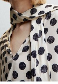 Femme portant un chemisier blanc à gros pois noirs et une écharpe assortie drapée autour du cou.