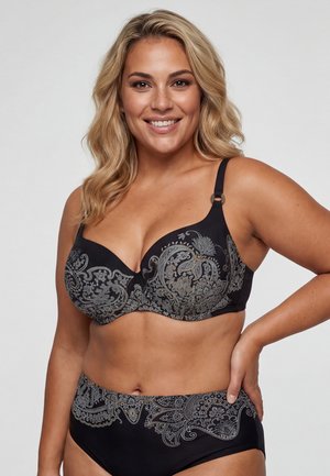 Uśmiechnięta kobieta z falującymi blond włosami, ubrana w dopasowany czarny biustonosz i majtki z wysokim stanem ozdobione misternymi srebrnymi wzorami paisley.