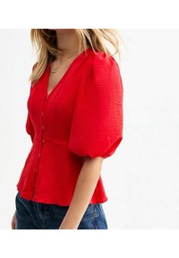 Rød bluse med pufærmer og V-udskæring, tekstureret stof og knaplukning foran. Kombination med blå denimjeans set fra siden.