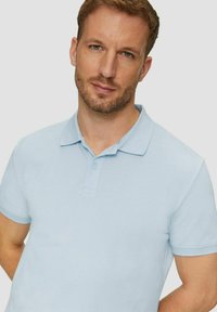 Mann mit leichtem Bart und blauen Augen, der ein hellblaues Polohemd mit kurzen Ärmeln trägt, steht vor einem schlichten, hellen Hintergrund.