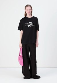 Black oversized t-shirt with a white graphic print, paired with loose black pants and a pink handbag for contrast. Smooth textures throughout.