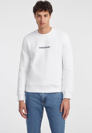Hvit crewneck hettegenser med "GUESS JEANS" logo i mørkeblå, ribbestrikkede mansjetter og hem, kombinert med lyseblå denimjeans.