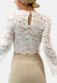 Weiße Spitzenbluse mit langen Ärmeln, im kurzen Design, mit gewelltem Saum und einem Schlüssellochausschnitt auf der Rückseite, der mit einem kleinen Knopf geschlossen wird.