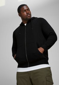 Black zip-up hoodie made of cotton blend, featuring front pockets, drawstring hood, and ribbed cuffs. Layered over a white T-shirt.