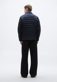 Granatowa kurtka puchowa z pionowym pikowanym wzorem, wysokim kołnierzem i bocznymi zamkami błyskawicznymi, zestawiona z szerokimi, ciemnymi spodniami dżinsowymi.