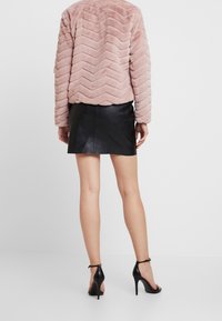 Kobieta w różowej kurtce z futra syntetycznego w wzór zygzak, czarnej skórzanej mini spódnicy i czarnych sandałach na wysokim obcasie, stojąca na białym tle.