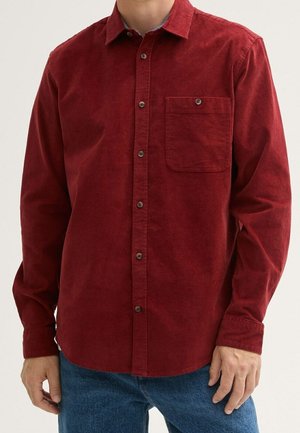 Mężczyzna ubrany w czerwoną sztruksową koszulę na guziki z długim rękawem i kieszenią z przodu oraz niebieskie dżinsy, stojący z rękami swobodnie opuszczonymi wzdłuż ciała.