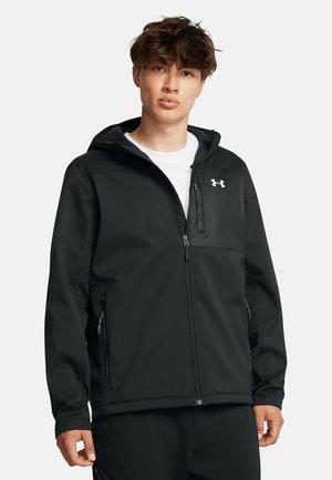 Giovane uomo con capelli ricci che indossa una felpa nera con zip Under Armour sopra una maglietta bianca su uno sfondo semplice.