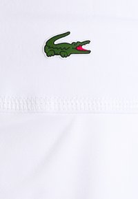 Bílá látka obsahuje zelené logo krokodýla s růžovými ústy. Design zahrnuje texturovaný, prošitý proužek podél spodního okraje.