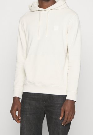 Felpa crema con tasca frontale, cappuccio regolabile con coulisse e polsini a coste. Presenta un piccolo logo sul petto. Indossata con jeans scuri.