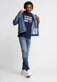 Dongerijakke, mørkeblå t-skjorte med hvite grafikker, lyseblå jeans og hvite joggesko. Modellen poserer med jakken åpen, og viser frem antrekket.