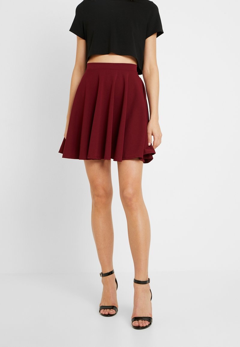 Femme portant un crop top noir, une jupe plissée bordeaux et des sandales à talons hauts noires avec bride à la cheville, debout devant un fond blanc uni.