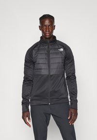 Chaqueta negra con un frontal acolchado, tela ligera, cuello alto y cierre de cremallera. Logo en el pecho; mangas ajustadas con orificios para los pulgares.