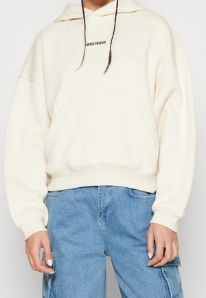 Person iført en off-white hættetrøje med teksten "WRSTBHVR" på brystet og løstsiddende lyseblå denim cargobukser.