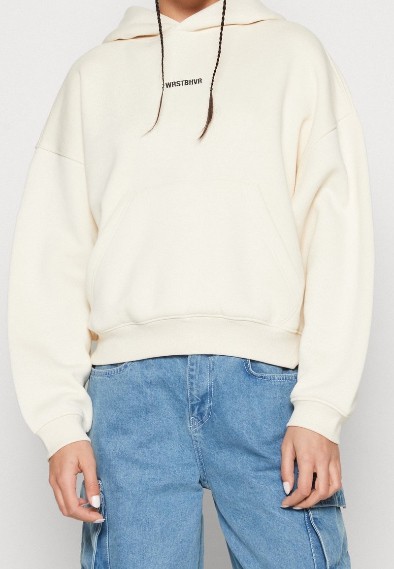 Personne portant un sweat à capuche écru avec le texte « WRSTBHVR » sur la poitrine et un pantalon cargo ample en jean bleu clair.