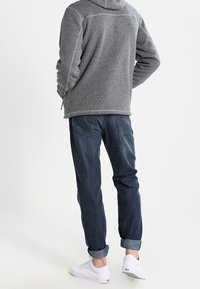 Szara polarowa bluza z kapturem, z tkaniny o fakturze, zestawiona z ciemnoniebieskimi dżinsami ze ściągaczami i białymi trampkami. Widok z tyłu.