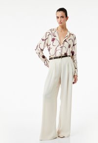 Losse witte broeken met plooien en een zwarte riem, gecombineerd met een crèmekleurige blouse met een maroon bloemenpatroon en een diepe halslijn.