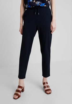 Mujer que lleva pantalones negros hasta el tobillo con cinturilla atada, una blusa floral y sandalias marrones de tacón, de pie sobre un suelo gris.