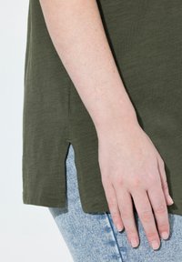 Oliivinvihreä, lyhythihainen toppi sivuaukkojen kanssa, valmistettu pehmeästä kankaasta. Yhdistetty vaaleansinisiin, kuluneisiin farkkuihin.