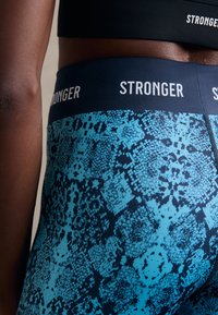 Siniset ja mustat legginsit, joissa on käärmeennahkakuosi, leveällä vyötärönauhalla, jossa lukee "STRONGER." Kangas näyttää sileältä ja joustavalta.