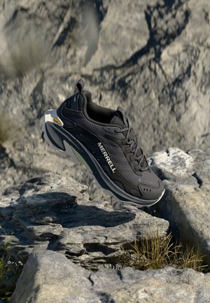 Czarny but trekkingowy Merrell z podeszwą Vibram unoszący się nad skalistym terenem z małymi roślinami i szarym kamiennym tłem.