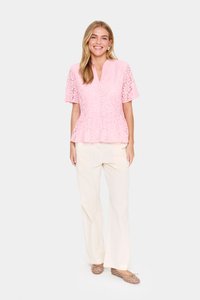 Haut en dentelle rose à manches courtes avec un design à basque, associé à un pantalon large crème et des ballerines marron à pois blancs.