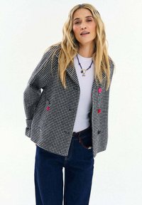 Žena s dlhými blond vlasmi, oblečená v čierno-bielom kockovanom saku s farebnými gombíkmi, bielom tričku, modrých džínsoch a náhrdelníku z korálok.
