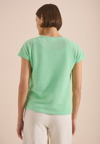 Femme aux cheveux courts et bruns portant un haut vert menthe à manches courtes perforé et un pantalon blanc, debout devant un fond beige.