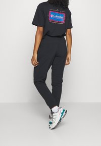 Svart t-shirt med kort ärm och färgglad Columbia Sportswear-logotyp på ryggen, i kombination med svarta avsmalnande byxor och träningssko.