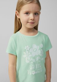 Mintgrünes Baumwoll-T-Shirt mit floralem Druck und erhabenen Akzenten. Text "Frühling Blumen" in Weiß am unteren Rand. Kurze Ärmel.
