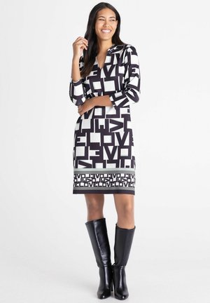 Lächelnde Frau steht mit der Hand nahe am Gesicht, trägt ein schwarz-weiß gemustertes Kleid mit dem Wort "LOVE" und schwarze kniehohe Stiefel.