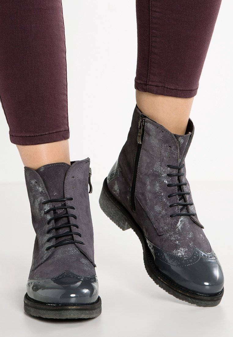 Personne portant des bottines à lacets en daim gris foncé et cuir brillant avec des fermetures éclair latérales et un pantalon violet foncé.