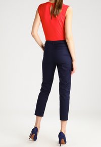 Röd ärmlös topp med en nyckelhålrygg, kombinerad med marinblå slim-fit byxor och blå högklackade skor, sedd bakifrån.