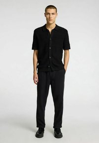 Camicia nera a maniche corte con bottoni, caratterizzata da un motivo testurizzato, abbinata a pantaloni neri larghi e scarpe nere testurizzate.