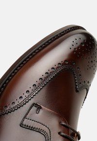 Scarpa elegante in pelle marrone con dettagli brogue, chiusura con lacci e design curvo. Presenta una finitura liscia con cuciture a contrasto e perforazioni.