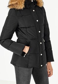 Veste noire rembourrée avec un col en fausse fourrure, des boutons-pression et des poches latérales zippées. Présente un design ajusté et une texture douce.