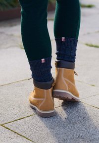 Botas de cuero marrón con acentos negros, costuras detalladas y una suela texturizada, combinadas con calentadores de piernas de canalé azul oscuro y pantalones verdes.