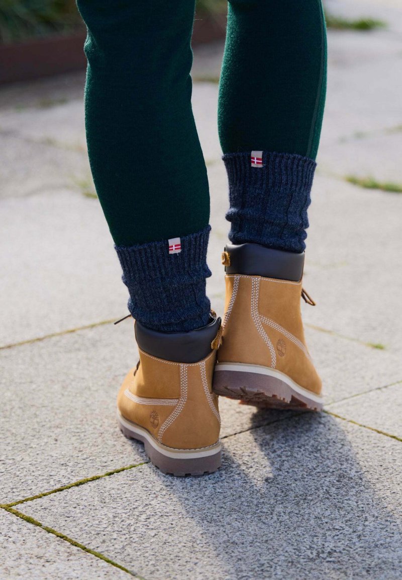 Botas de cuero marrón con acentos negros, costuras detalladas y una suela texturizada, combinadas con calentadores de piernas de canalé azul oscuro y pantalones verdes.
