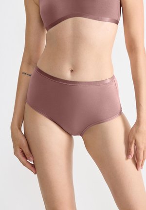 Vrouw in een stofroze naadloze high-waist shorts en bijpassende beha, staande met ontspannen armen tegen een effen witte achtergrond.