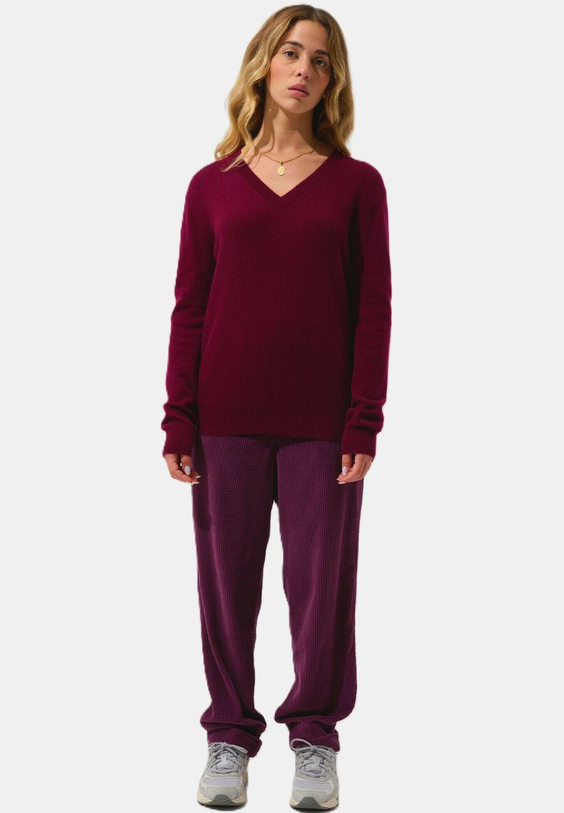 Burgundyröd V-ringad tröja i mjukt garn, kombinerad med strukturerade lila byxor. Modellen bär grå sneakers, med avslappnade händer vid sidorna.