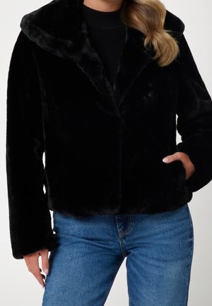 Femme portant une veste en fausse fourrure noire à large col, une main dans la poche, associée à un jean bleu et un haut noir côtelé.
