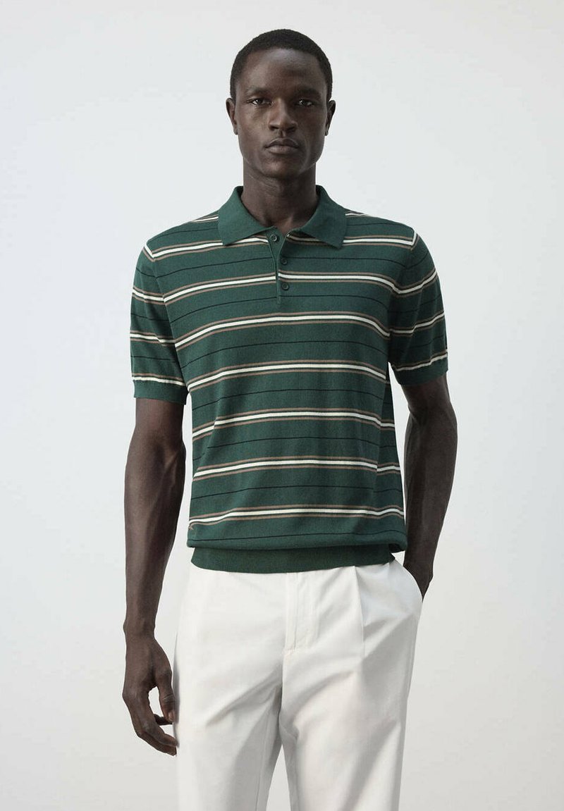 Homme portant un polo vert rayé à manches courtes rentré dans un pantalon blanc, sur un fond clair uni.