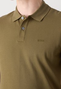 Polo vert olive avec un tissu texturé, deux boutons argentés et un logo "BOSS" subtilement embossé sur la poitrine.