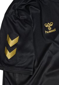 Maglietta sportiva nera con maniche corte, con il logo "hummel" dorato e due motivi a chevron dorati sulla manica sinistra. Tessuto liscio.