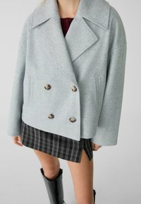 Manteau croisé gris clair en mélange de laine avec grands cols et boutons en écaille, associé à une mini-jupe noire à carreaux et des bottes noires brillantes.